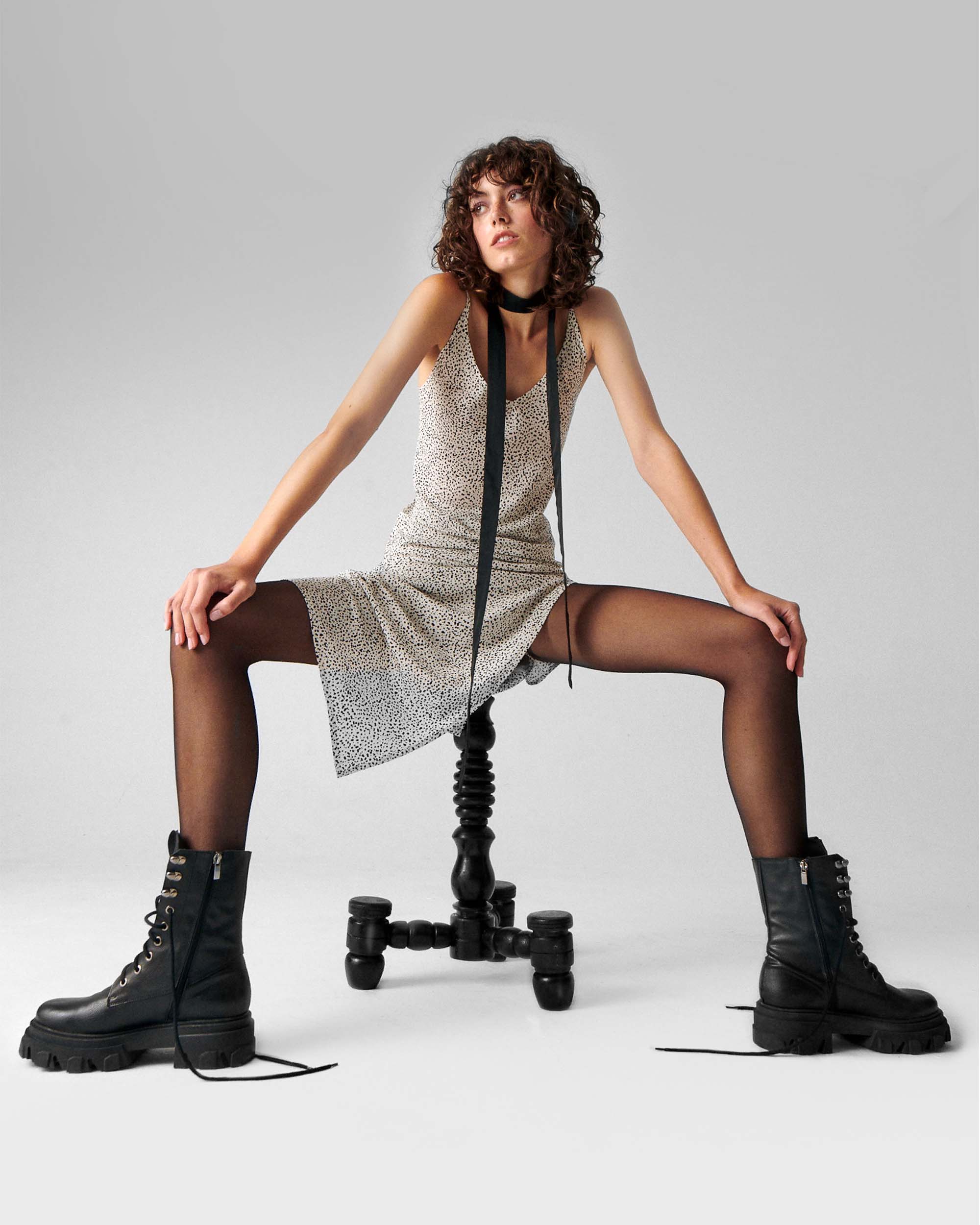 Woman in black combat boots, patterned dress, and black tights sitting on modern stool